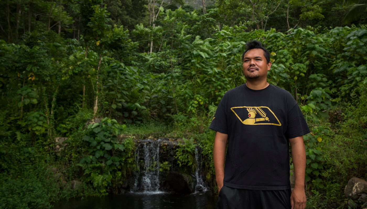 A man stands in front of a lush jungle waterfall, surrounded by greenery