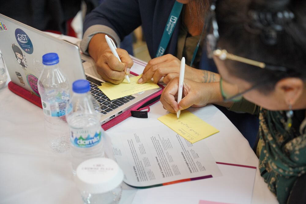 Two women at a table, writing in notebooks with pens